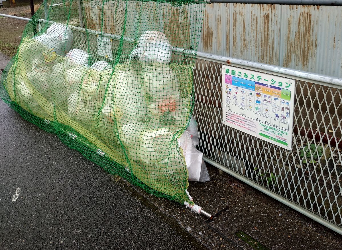 幸手市の集積所。建物の横の道路沿いに、黄色い指定袋がネットをかけて並んでいる